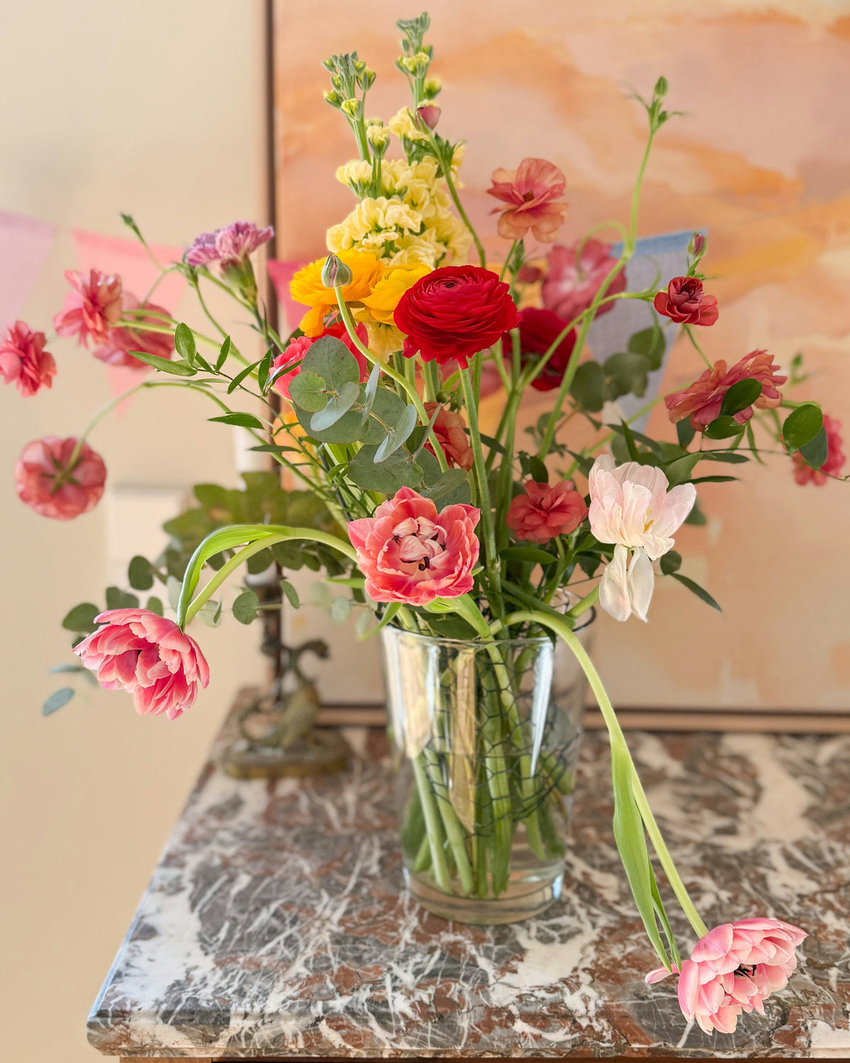Pink ranunculus, tulips and yellow stocks in a glass vase on a marble table